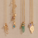 Four gold necklaces with unique charms are displayed on a beige fabric wall. The necklaces feature a dried flower, a blue gemstone, a green crystal, and a gold leaf pendant with a purple heart gemstone and clear stones.