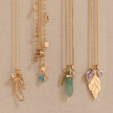 Four gold necklaces with unique charms are displayed on a beige fabric wall. The necklaces feature a dried flower, a blue gemstone, a green crystal, and a gold leaf pendant with a purple heart gemstone and clear stones.