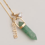 A gold-tone chain necklace featuring a green pointed crystal pendant, a small clear heart-shaped gemstone, and a vertical gold nameplate charm spelling 'emilia', displayed on a light gray background.