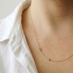 A delicate gold chain necklace worn by a model, featuring a small openwork letter 'B' pendant and a tiny pink gemstone accent, styled with a white linen shirt.