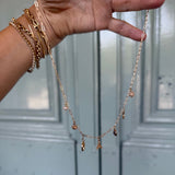 Gold necklace with small pendants held by a hand against a light blue paneled wall.