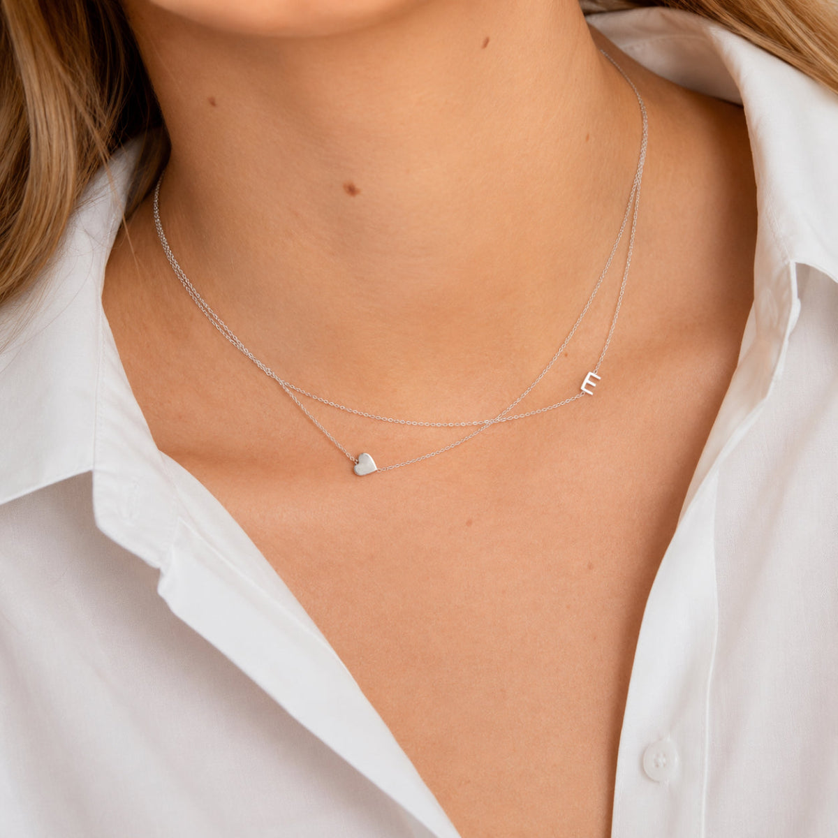 Close-up of a person wearing a silver necklace with a heart charm and letter 'E' charm on a white shirt background.