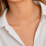 Close-up of a person wearing a silver necklace with a heart charm and letter 'E' charm on a white shirt background.