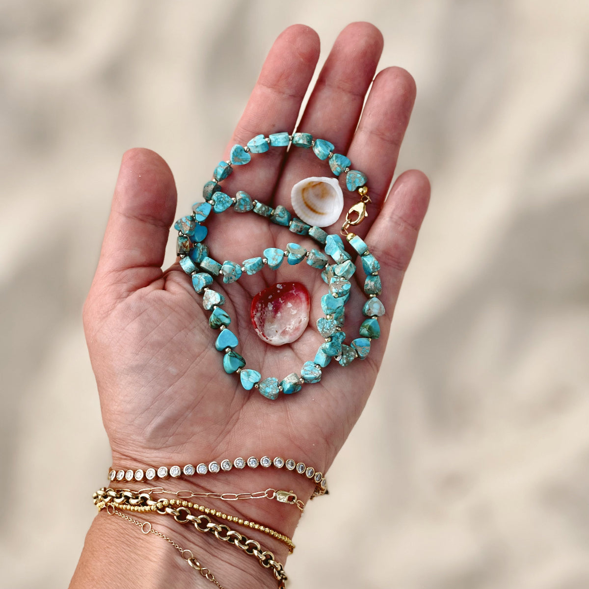 A hand holding an Endless Summer Necklace - Sea + Sky with two shells, and wearing several bracelets, including a Love Set Match Bracelet, a Diamond Cut Bracelet, a Rolo Bracelet and an Element Short Link Bracelet with a blurred sand background. 