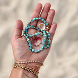 A hand holding an Endless Summer Necklace - Sea + Sky with two shells, and wearing several bracelets, including a Love Set Match Bracelet, a Diamond Cut Bracelet, a Rolo Bracelet and an Element Short Link Bracelet with a blurred sand background. 