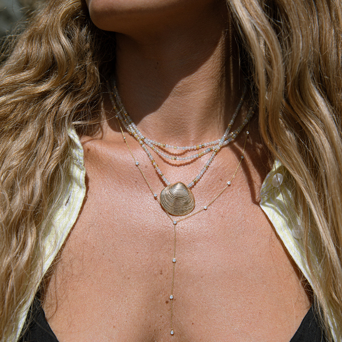 Close-up of a woman’s décolletage wearing Endless Summer Necklaces – Saint Tropez, including one with The Golden Venus Shell, and a Starstruck Diamond Lariat Necklace, styled in a light yellow and white striped shirt with blonde hair.