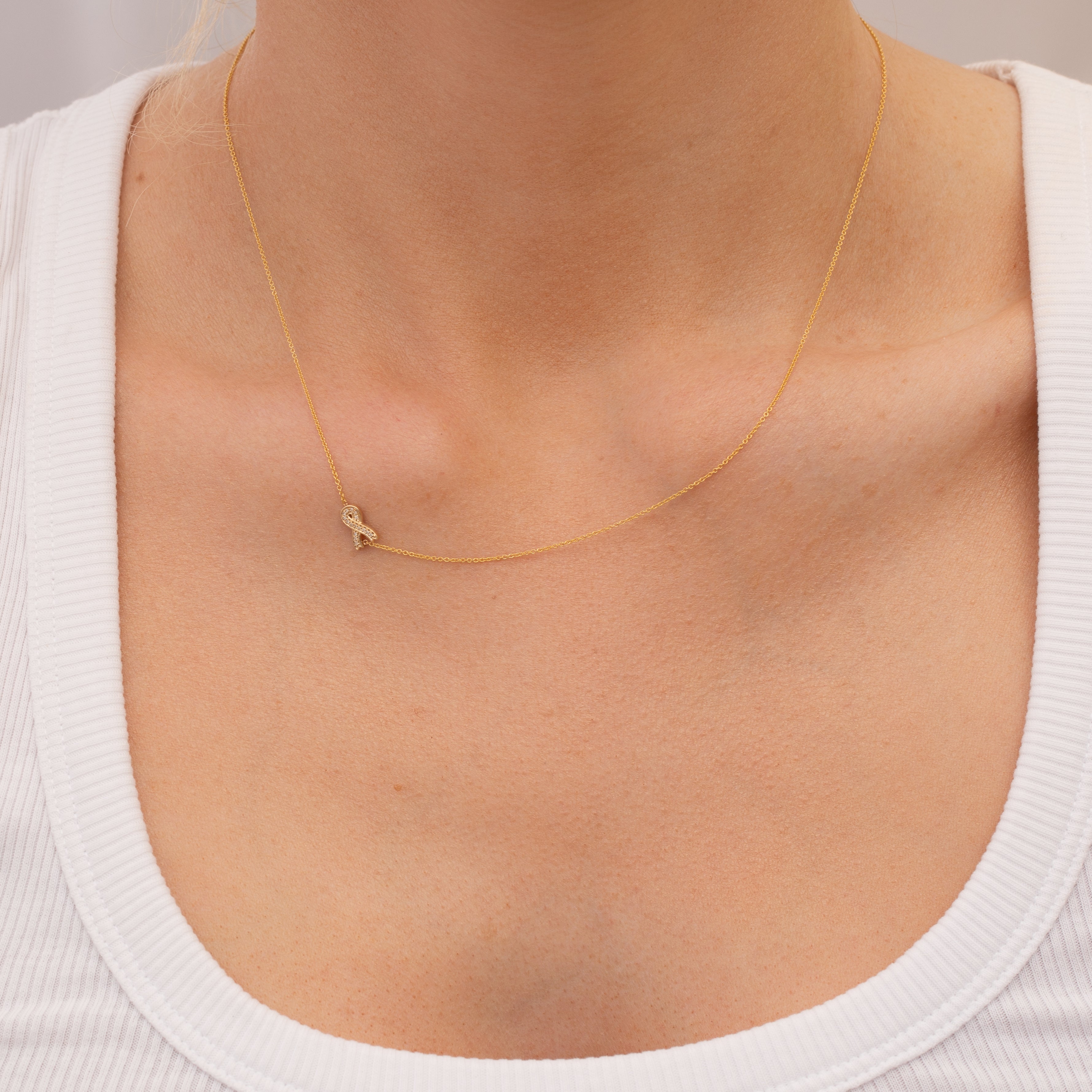 Gold necklace with a small pavé diamond ribbon charm on a person wearing a white top.