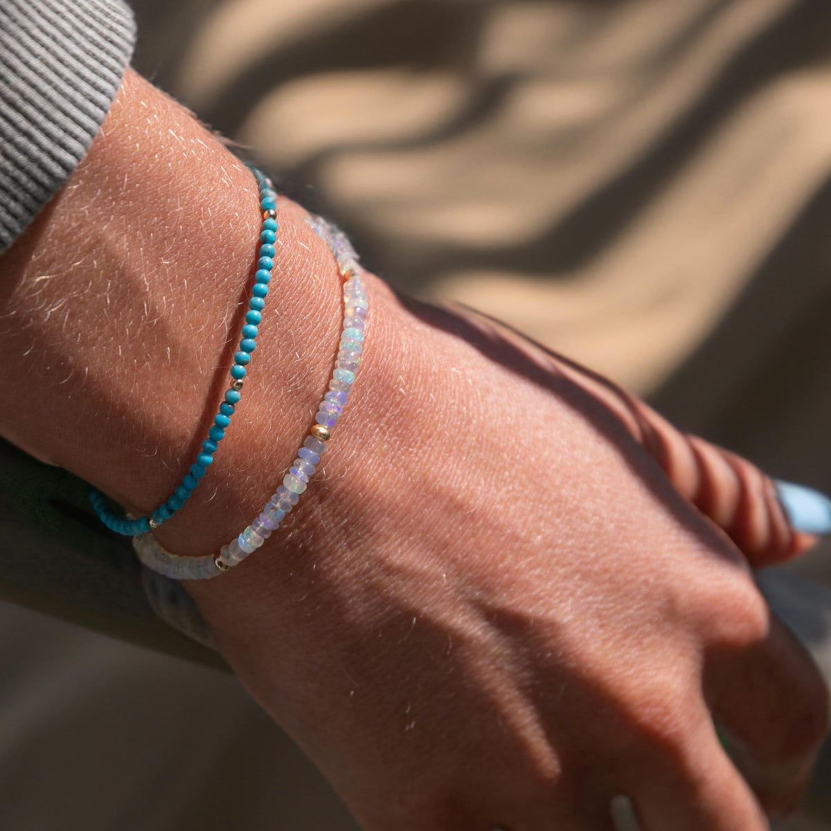 Close-up of a handing wearing two Endless Summer Bracelets (Turqs + Caicos and Blue Lagoon) with a blurred beige background. 