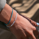 Close-up of a handing wearing two Endless Summer Bracelets (Turqs + Caicos and Blue Lagoon) with a blurred beige background. 