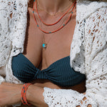 Close-up of a person’s décolletage and arm wearing an Endless Summer Bracelet and Necklace in Mykonos, an Endless Summer Necklace (including one wrapped around the wrist), a Classic Bead Chain with a Petite Nameplate Pendant, an Etched Turquoise Heart Charm and a Diamond Pendant, styled with a navy blue bikini top and a white crochet top.