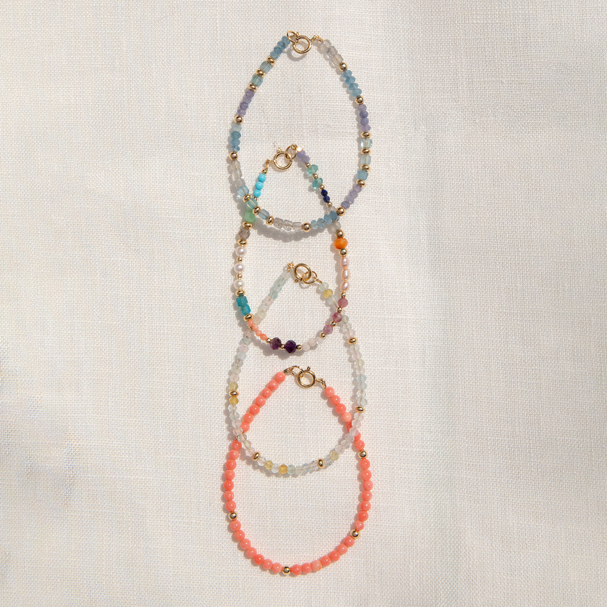 Four Endless Summer Bracelets (Blue Lagoon, Capri, Mykonos and Saint-Tropez) laid vertically on an off-white fabric background. 