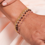 A close-up of a wrist wearing three Bouquet Bracelets (grey, maroon and pink) and two Bubble Bracelets in yellow gold. 
