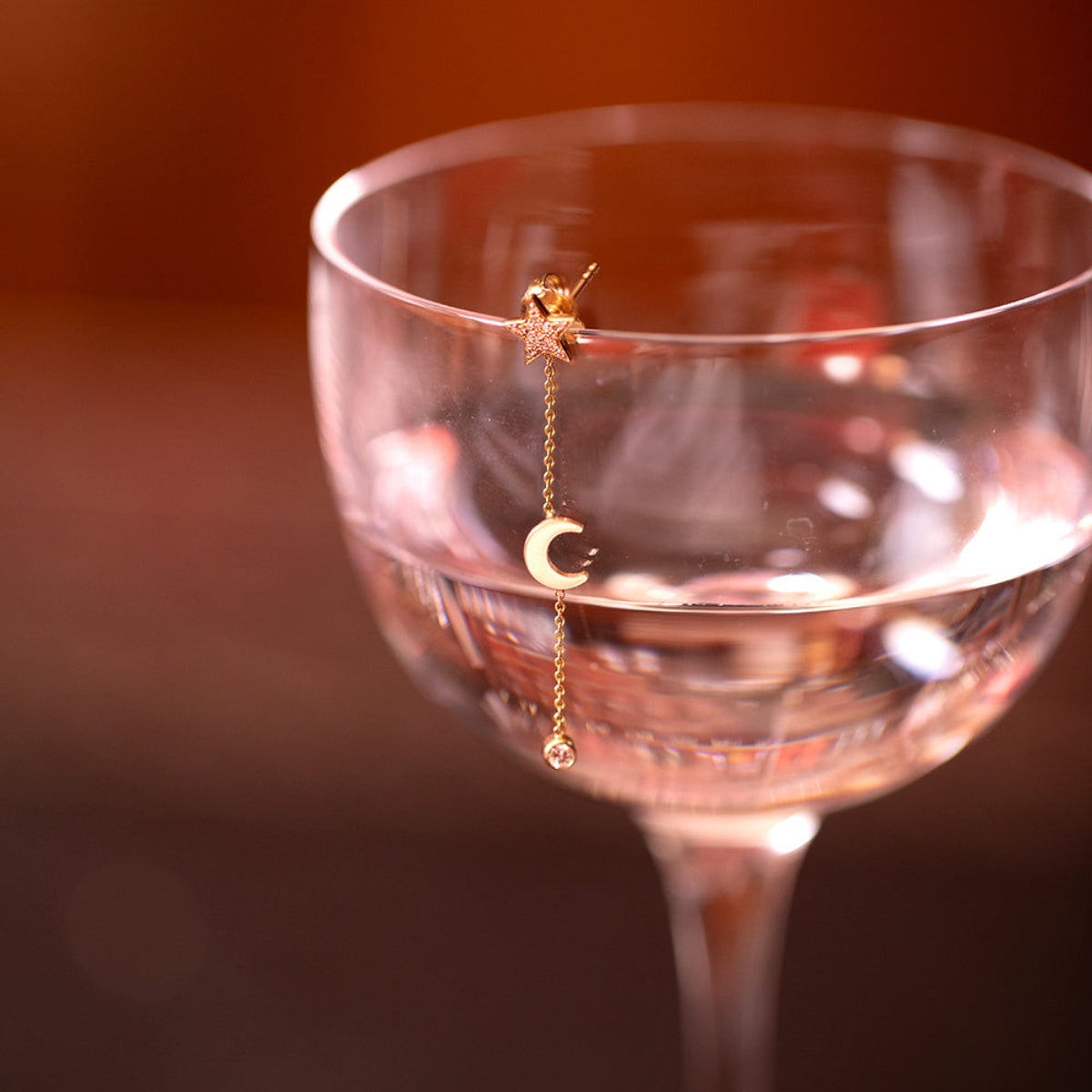 A Luna Spritz Earring on top of a cocktail glass against a blurred red background.
