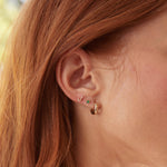 Close-up of a 14K Gold Pavé Letter Stud in J, a Birthstone Stud - Emerald (May), and a Gold Huggie worn in a woman’s ear, with orange hair visible.