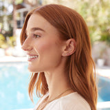 A woman wearing two Birthstone Studs, a Diamond Bar Earring, a Box Chain,and a Quintet Birthstone Layering Necklace in Turquoise, looking to the side with orange hair and a white top, with a blurred outdoor pool background.