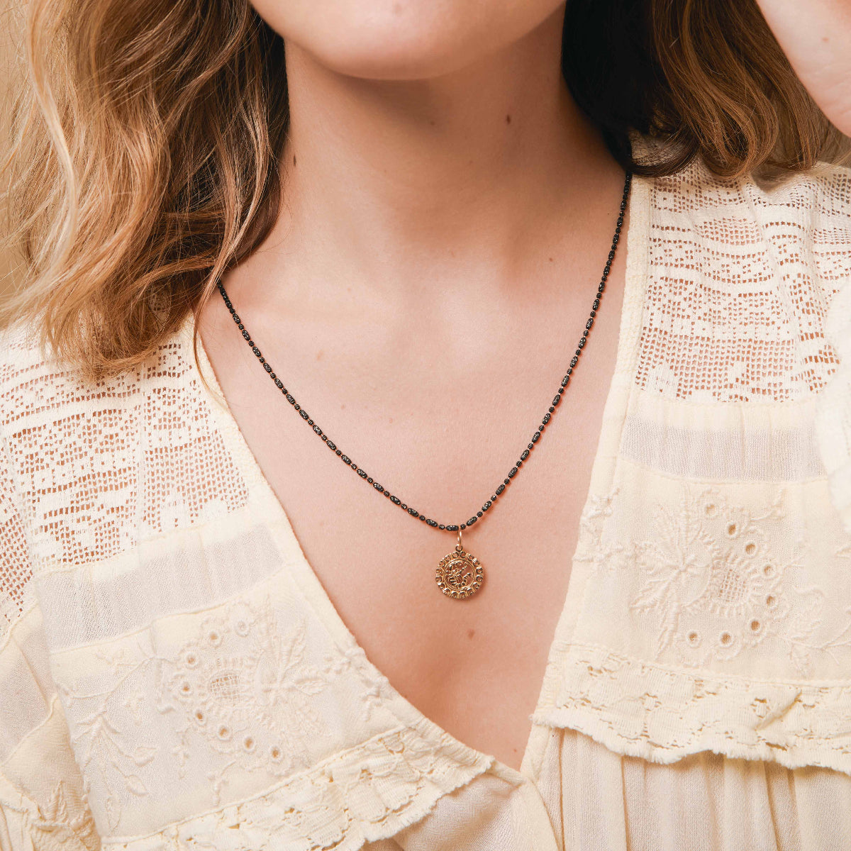 A close-up of a blonde woman's décolletage wearing a Black Rhodium Antique Bead Chain with a Zodiac Pendant  and a light yellow lace top. 