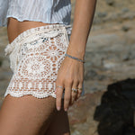 A close-up of a person’s arm wearing two Endless Summer Bracelets (Blue Lagoon), The Forget Me Not Bouquet Ring, a Tres Pearl Ring and an Open Diamond Ring, styled in cream lace shorts and a light blue top, with a blurred stone background.