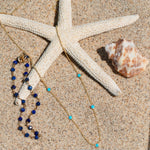 The Thistle Bouquet Bracelet and Quintet Birthstone Layering Necklace in yellow gold with turquoise stones, both laid in front of a starfish next to a shell against a sandy background.