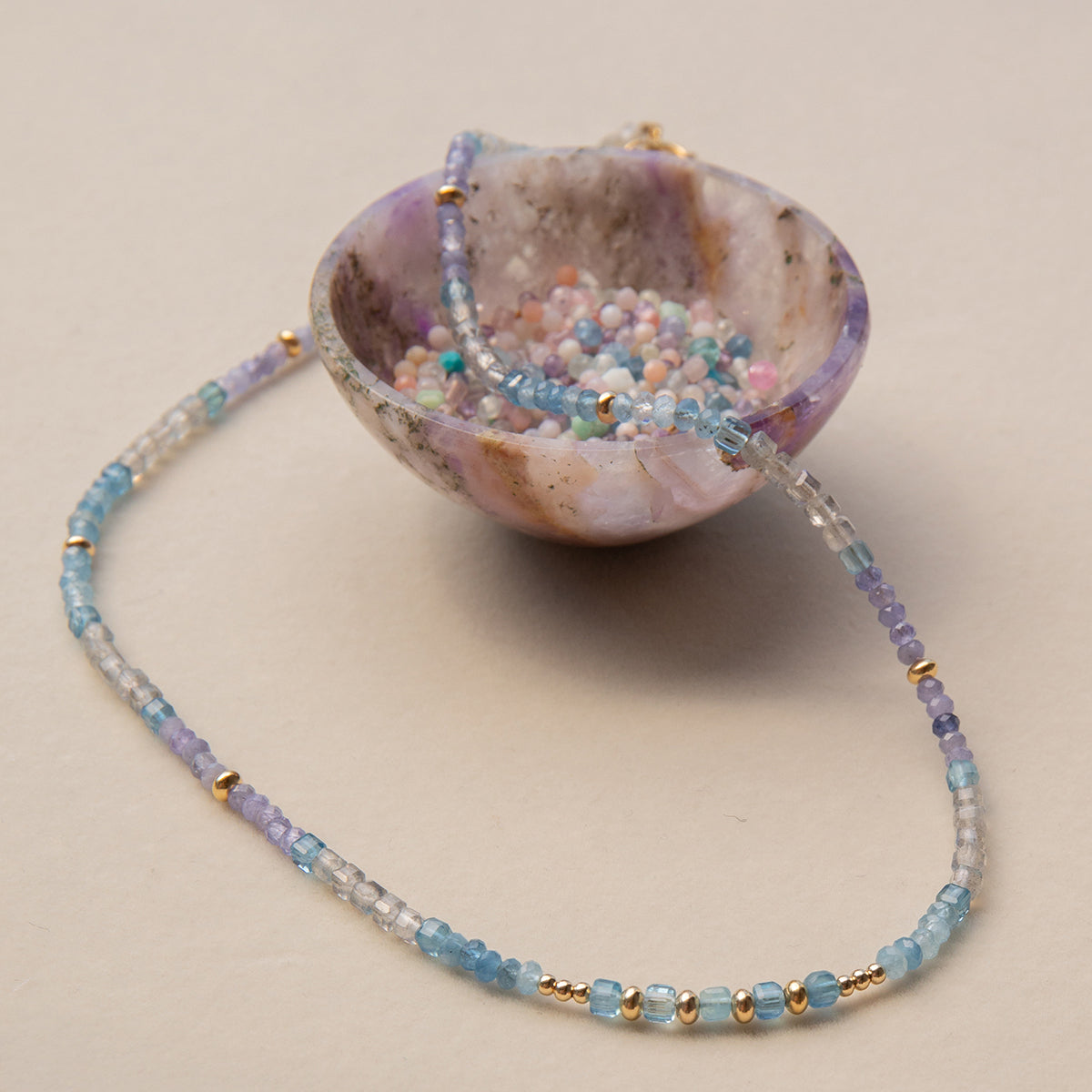 An Endless Summer Necklace - Blue Lagoon laid on top of a small coloured bowl with different coloured beads in it, against a light background.