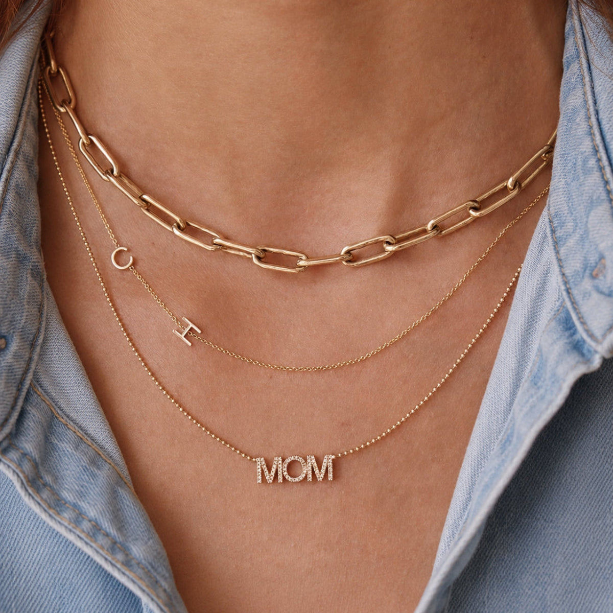 Gold necklaces with 'MOM' pendant and chain necklace on a person wearing a denim shirt.