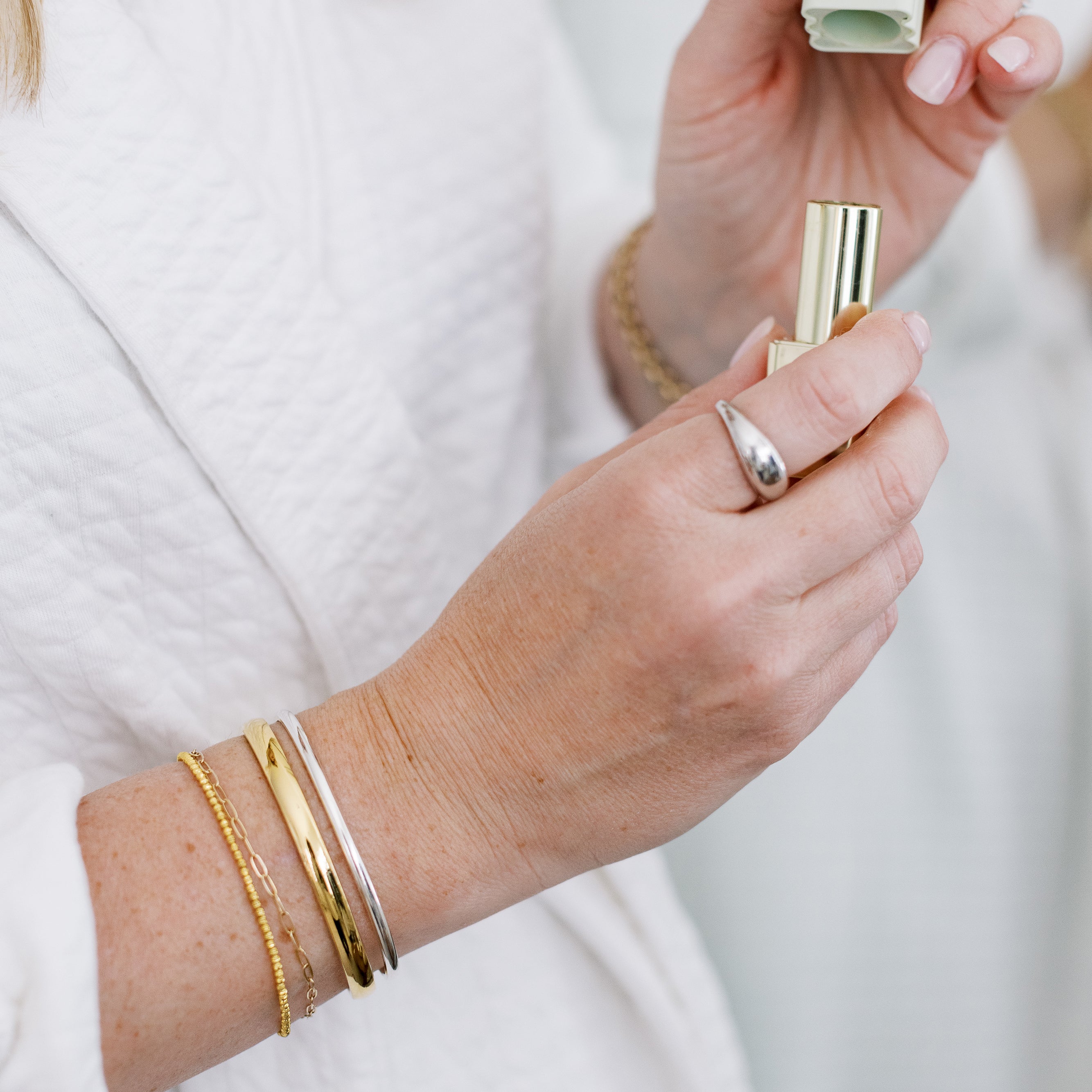 Close-up of a wrist wearing four gold bracelets holding a gold lipstick with a blurred background