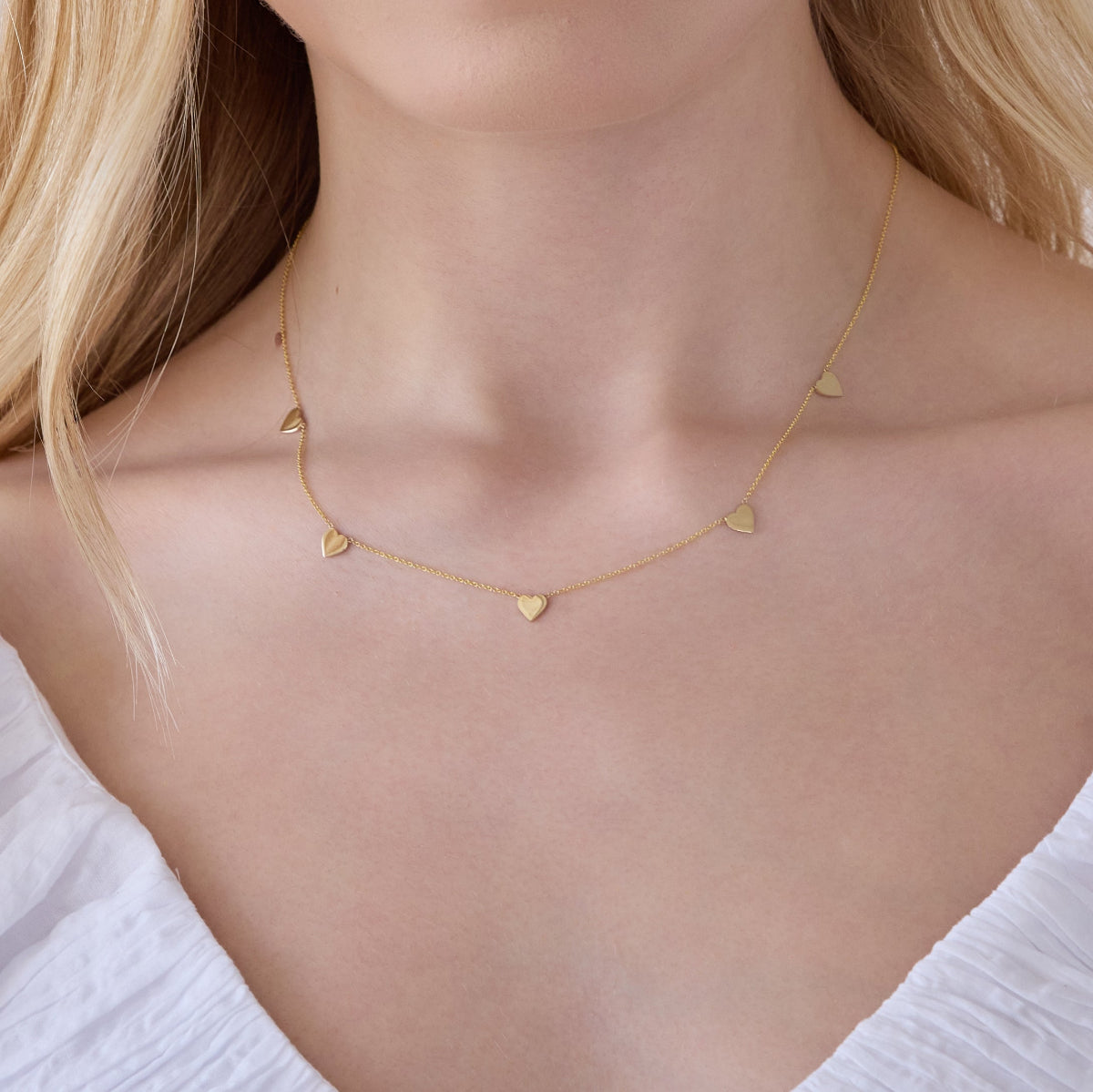 Close up of a  woman's neck wearing a 5 of Hearts Necklace in yellow gold with a white top and her blonde hair partially visible. 