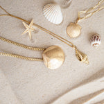 A Chic Cuban Curb Chain with The Golden Venus Shell, Element Long Link Chain with a Golden Scallop Shell and a Golden Spiral Shell on The Slide Chain against a sandy background. 