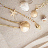 A Chic Cuban Curb Chain with The Golden Venus Shell, Element Long Link Chain with a Golden Scallop Shell and a Golden Spiral Shell on The Slide Chain against a sandy background. 