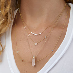 A close-up of a woman’s neck wearing four yellow gold necklaces: two Custom Gold Necklaces (one with three charms and the other with four charms), the Clarity Retreat Necklace and the Diamond Arc Layering Necklace, with a white top.