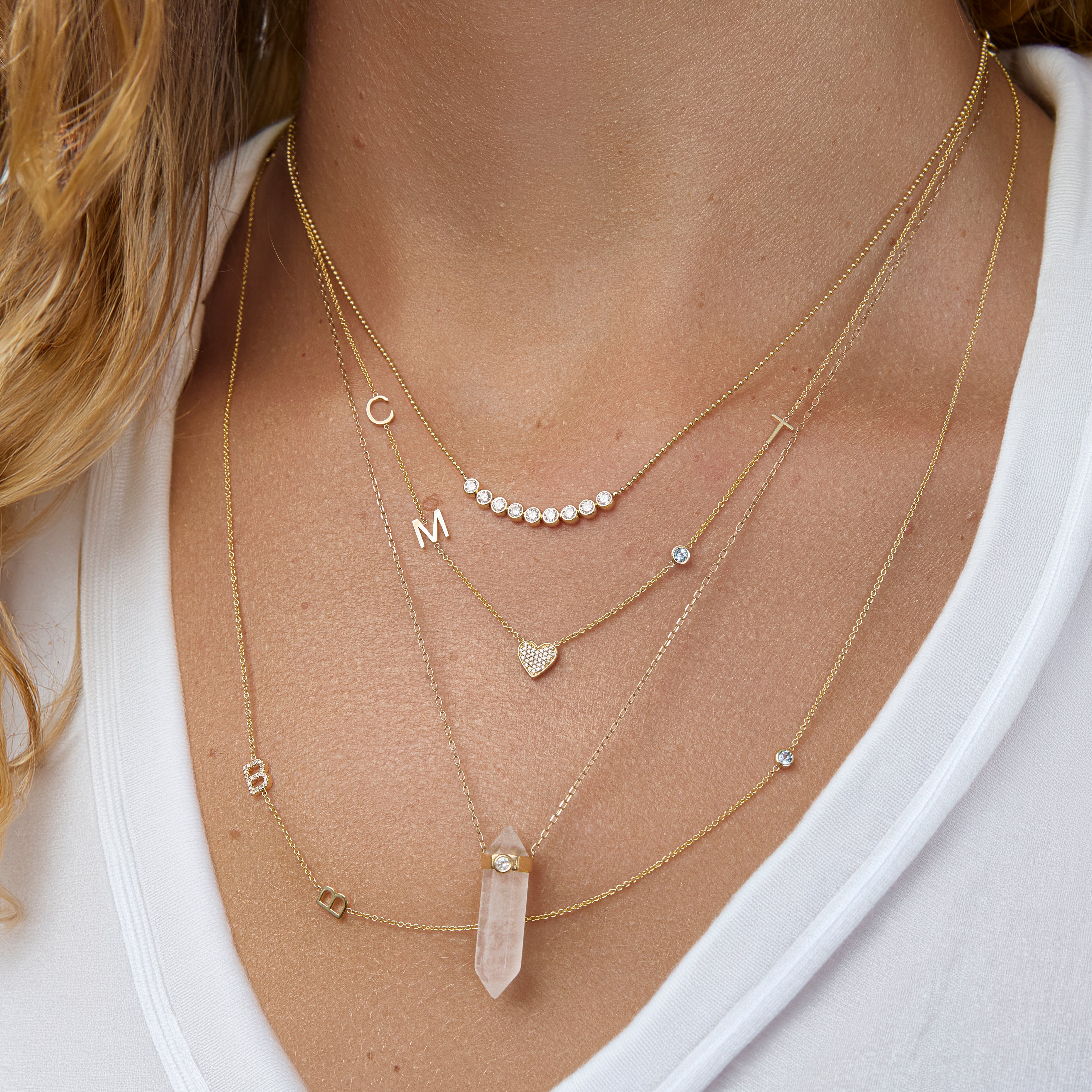 A close-up of a woman’s neck wearing four yellow gold necklaces: two Custom Gold Necklaces (one with three charms and the other with four charms), the Clarity Retreat Necklace and the Diamond Arc Layering Necklace, with a white top.