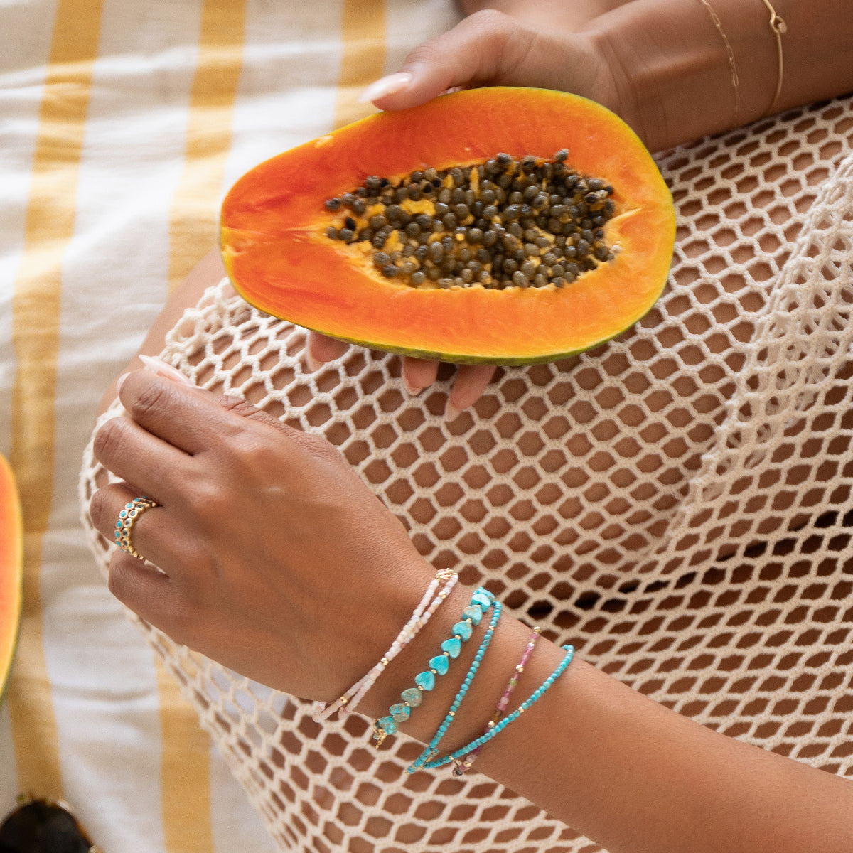 A hand wearing several Endless Summer Bracelets (Pink Sands, Turqs + Caicos, Coral Reef and Sea + Sky) and two Bouquet Rings, holding a papaya in the other hand with an Element Short Link Bracelet, styled in a cream mesh dress with a white and yellow striped background.