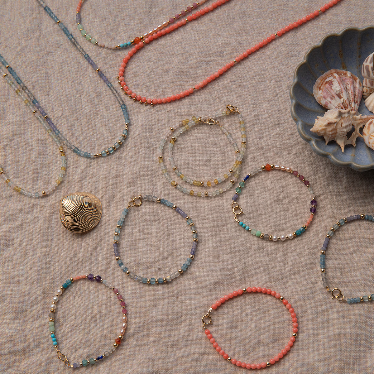 A range of Endless Summer Necklaces and Bracelets (Blue Lagoon, Mykonos, Saint-Tropez and Capri) laid on a beige fabric background with shells in a blue shell bowl. 