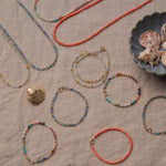 A range of Endless Summer Necklaces and Bracelets (Blue Lagoon, Mykonos, Saint-Tropez and Capri) laid on a beige fabric background with shells in a blue shell bowl. 
