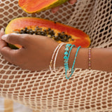 A hand wearing several Endless Summer Bracelets (Pink Sands, Turqs + Caicos, Coral Reef and Sea + Sky), holding a papaya while wearing cream mesh clothing.