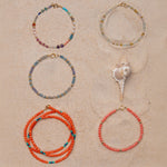 Four Endless Summer Bracelets (Capri, Saint-Tropez, Blue Lagoon and Mykonos) and an Endless Summer Necklace (Calypso) on a sandy background with a shell. 