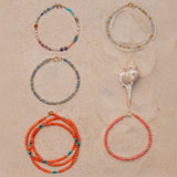 Four Endless Summer Bracelets (Capri, Saint-Tropez, Blue Lagoon and Mykonos) and an Endless Summer Necklace (Calypso) on a sandy background with a shell. 