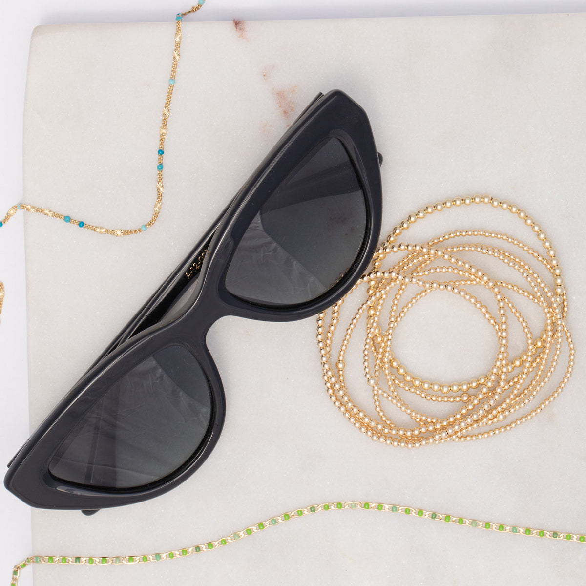 Two Confetti Necklaces in blue and green, bubble bracelets and black sunglasses on a light marble surface. 