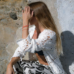 A woman kneeling down wearing Endless Summer Necklaces in Calypso and Mykonos (including one wrapped around the wrist) and several rings including a Tres Pearl Ring, styled with a navy blue bikini top, a white crochet top and black and white patterned bottoms against a stone background.