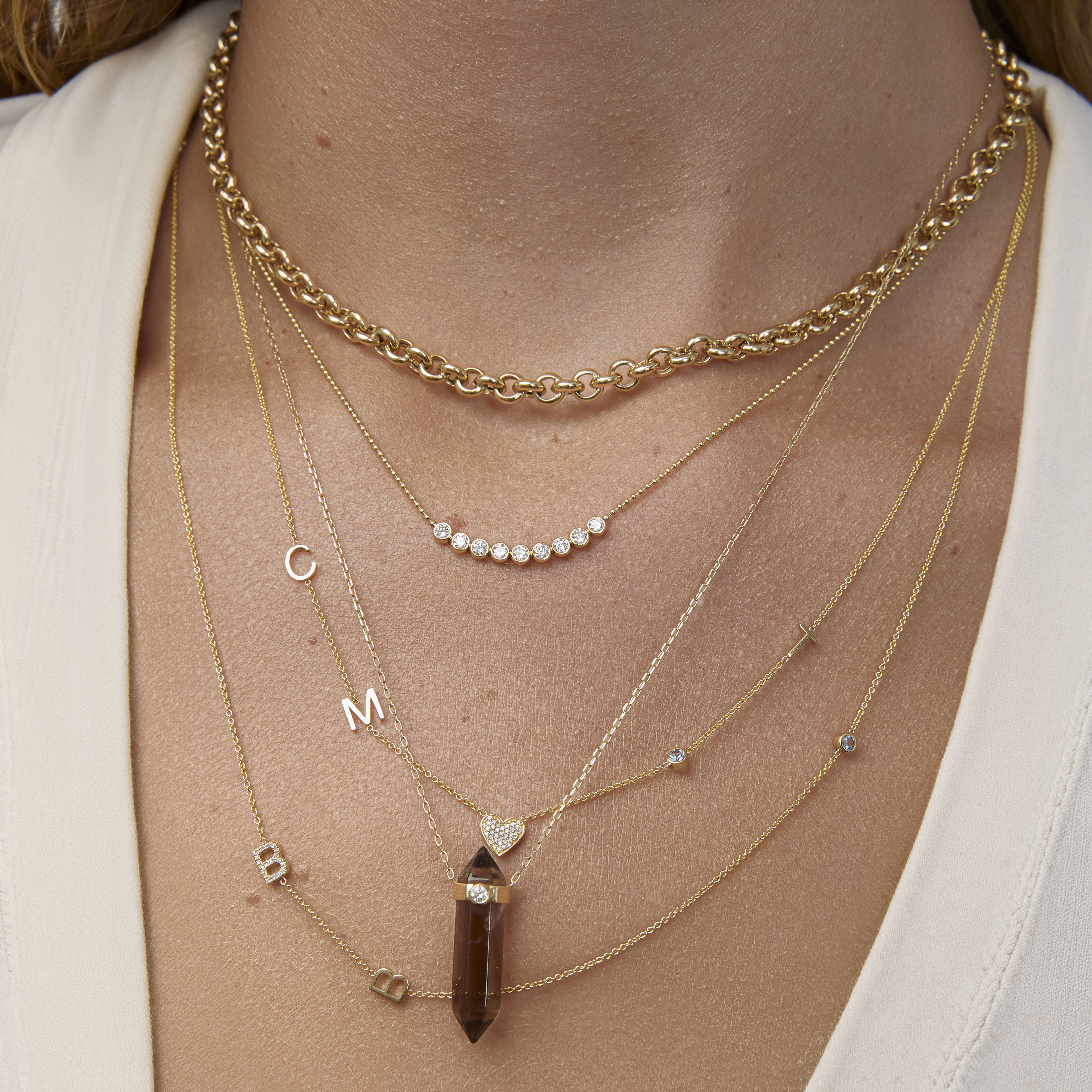 Close-up of a person wearing multiple gold necklaces with various pendants on a neutral background.