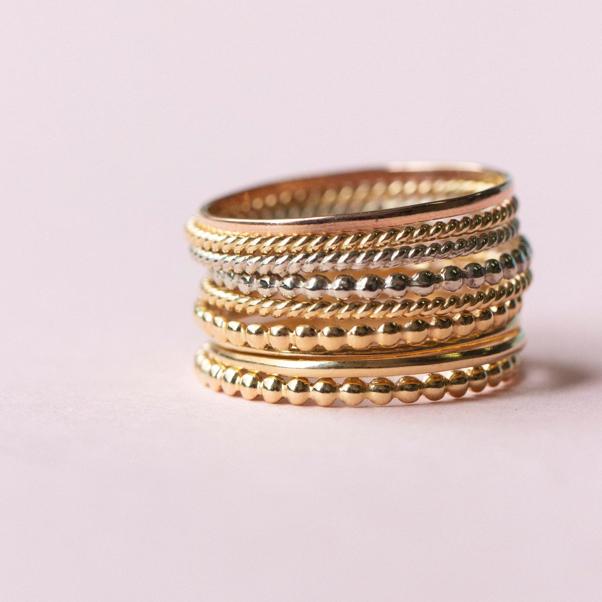 Several Delicate Trio Stacking Rings in yellow gold and white gold on a light pink background.