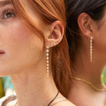 Close-up of a woman wearing a Diamond Cascade Earring in yellow gold and two Birthstone studs (diamond and pearl) with orange hair, with another woman’s ear in the background blurred, also wearing a Diamond Cascade Earring.