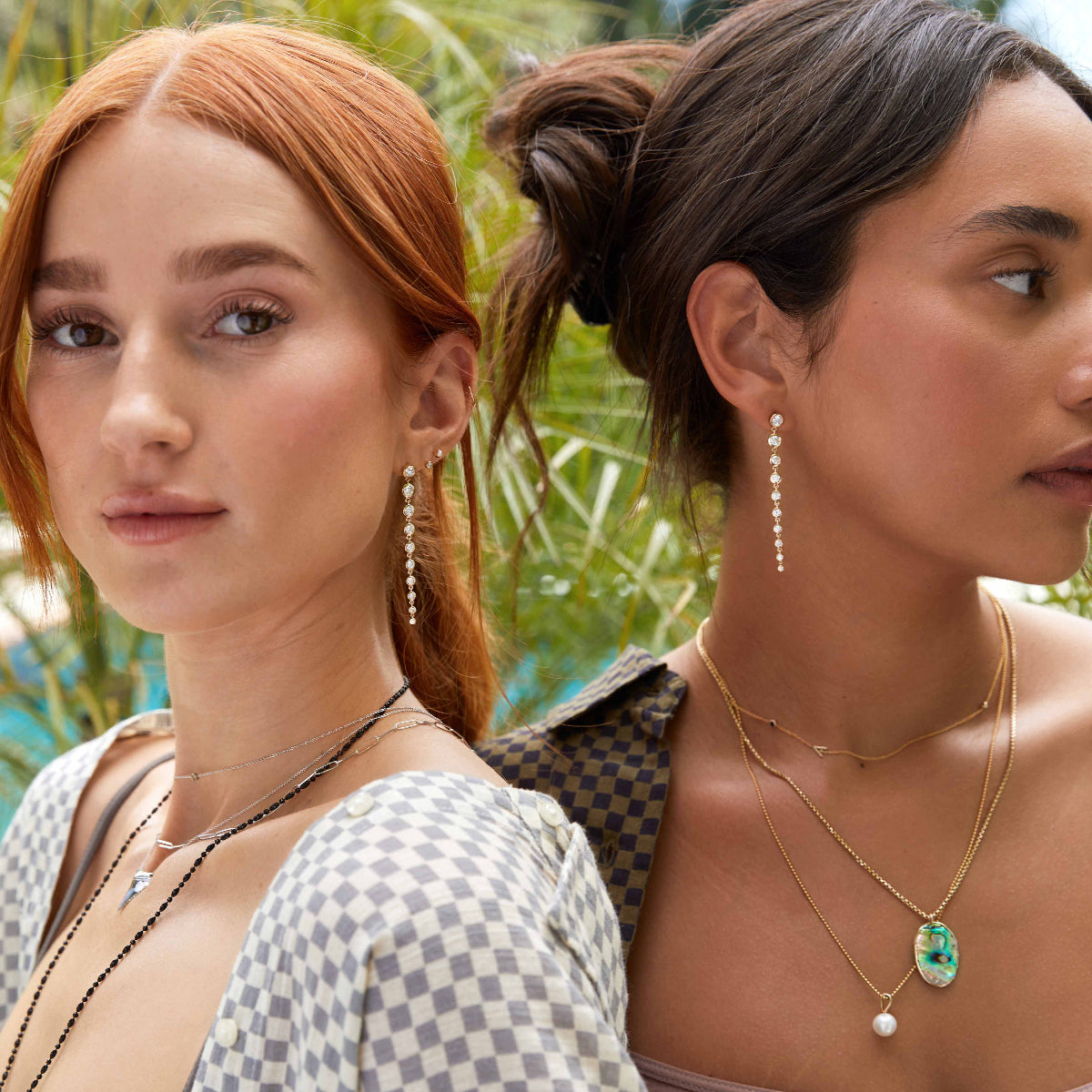 Two women wearing a Diamond Cascade Earring each, along with several necklaces including a Black Rhodium Antique Bead Chain, an Element Long Link Chain, a Perla Necklace and a Box Chain with a Large Abalone Cabochon Charm, styled in checkered tops with plants in the background.