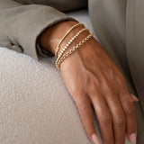 A close-up of a woman’s hand wearing an Element Short Link Bracelet, a Down the Line Bracelet, a Love Set Match Bracelet and a Rolo Bracelet, all in yellow gold, styled in a sage green jacket against a neutral background.