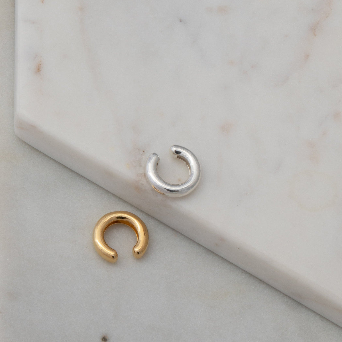 Two Ready to Mingle Ear Cuffs, one in sterling silver and the other in gold vermeil on a white marble surface.