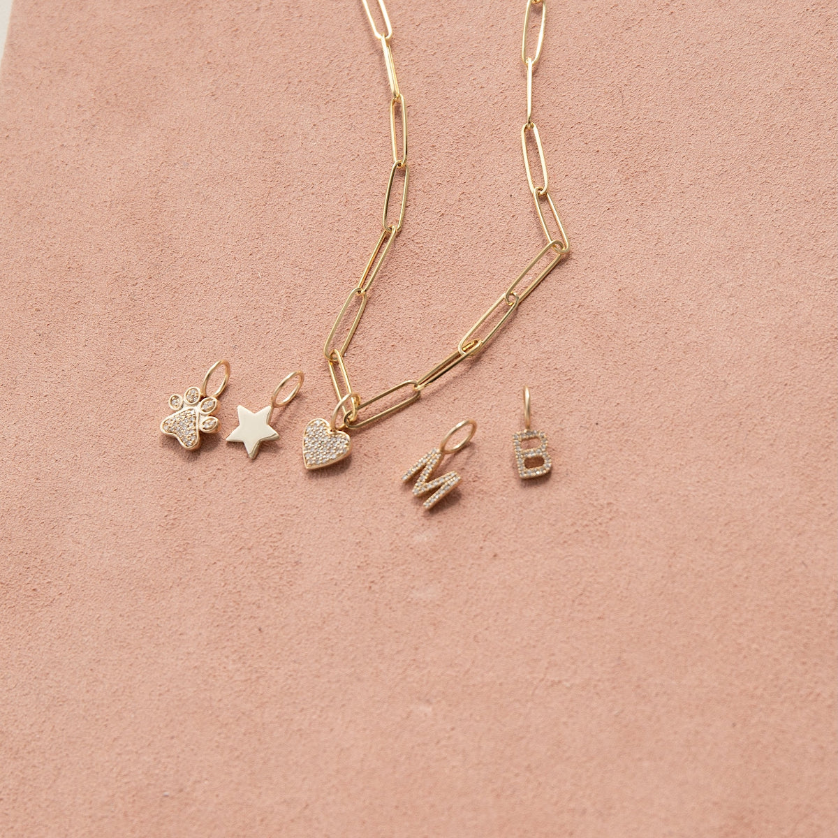 An Element Long Link Chain with Charm Pendants Star, Pave Paw and Pave Heart, and Pave Letter Charms in M and B, against a light pink background. 