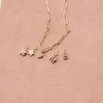 An Element Long Link Chain with Charm Pendants Star, Pave Paw and Pave Heart, and Pave Letter Charms in M and B, against a light pink background. 