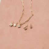 An Element Long Link Chain with Charm Pendants Star, Pave Paw and Pave Heart, and Pave Letter Charms in M and B, against a light pink background. 
