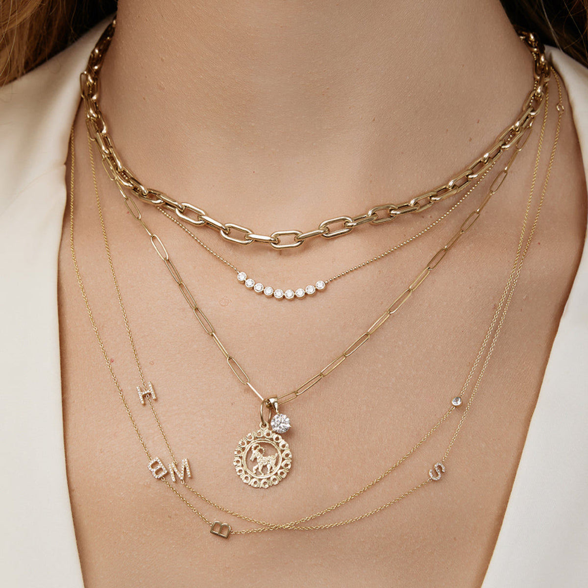 A close-up of a neck wearing a Bold Link Chain, a Diamond Arc Layering Necklace, an Element Long Link Chain with a Diamond Pendant and Zodiac Pendant (Aries) and two Custom Gold Necklaces - 3 Letters, all in yellow gold, styled with a cream top. 