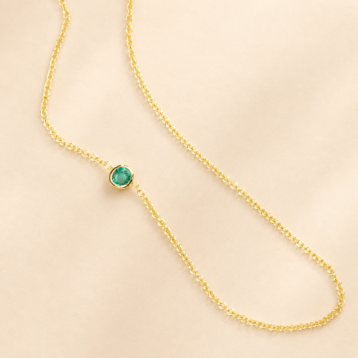 A yellow gold 14K Gold Asymmetrical Birthstone Necklace - Emerald (May) on a beige background.
