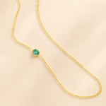 A yellow gold 14K Gold Asymmetrical Birthstone Necklace - Emerald (May) on a beige background.